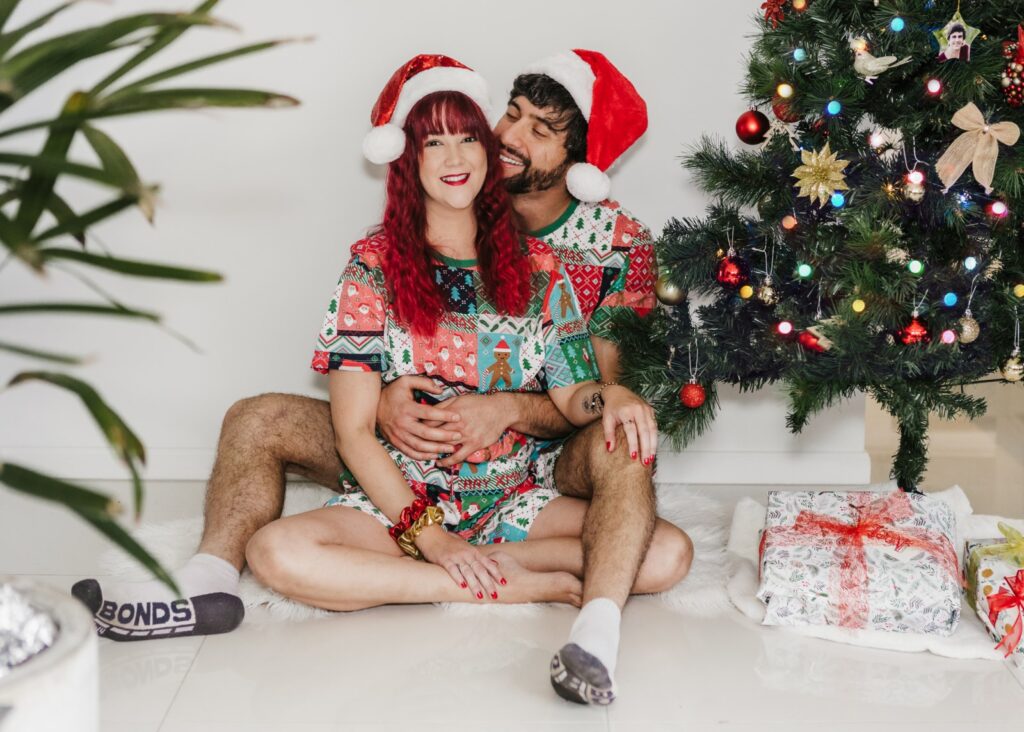 Couple wearing matching christmas pajamas and santa hats cuddling by the Christmas tree an an in home family session